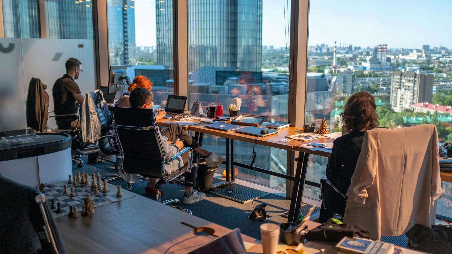 Open-plan office on a high floor with people working at laptops beside floor-to-ceiling windows overlooking city towers, with a chessboard on a nearby table